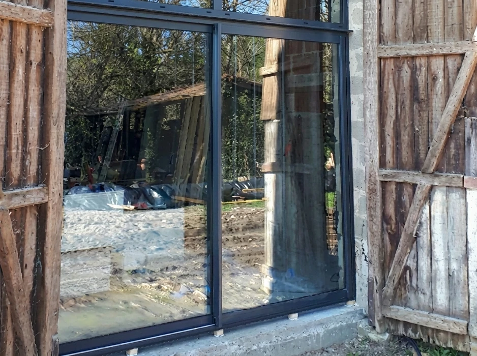Chantier de rénovation extérieur, vue sur une baie vitrée coulissante aluminium à double vitrage, encadrée de colombages en bois ancien et de parpaings