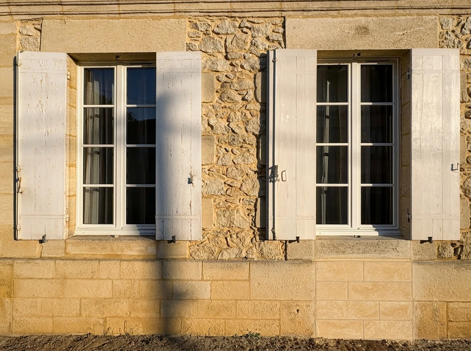 Façade en pierre calcaire avec deux fenêtres à croisillons, encadrements blancs, équipées de volets battants bois à deux vantaux, peints en blanc cassé, fixés par pentures métalliques noires