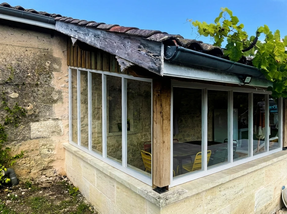 Véranda adossée à une maison en pierre calcaire, structure aluminium blanc, vitrages fixes et coulissants, poteaux en bois massif vieilli