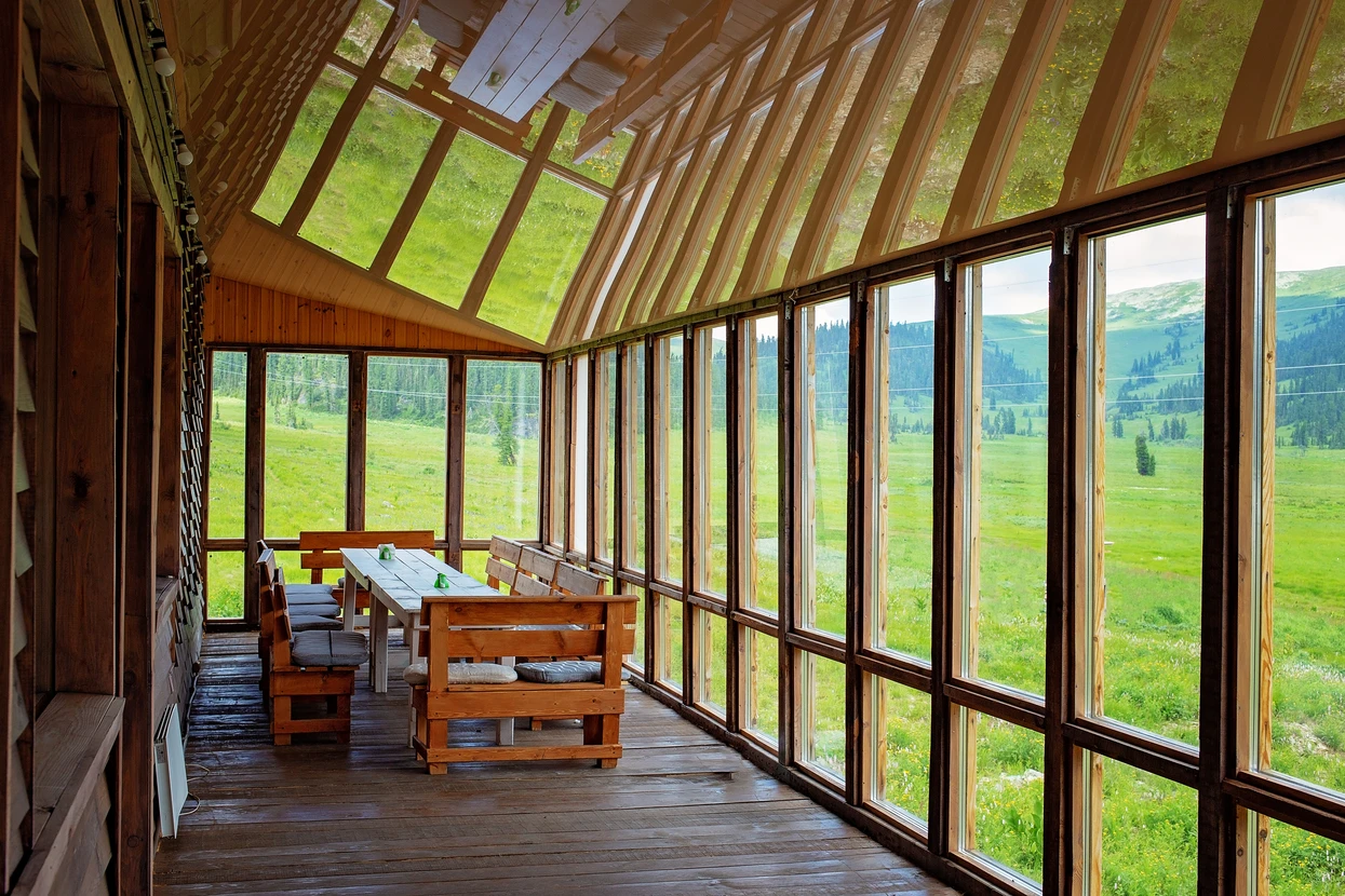 Véranda en bois avec structure apparente, grandes baies vitrées panoramiques donnant sur prairie verdoyante et forêt de conifères
